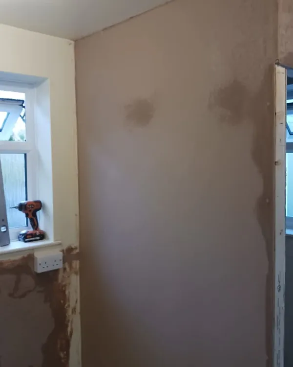 Freshly plastered wall with darker drying patches, beside a window with a spirit level and drill on the sill